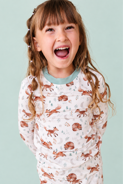 A young girl with long light brown hair wearing a long-sleeve bamboo pajama top with a woodland print of foxes, squirrels, and trees on a cream background, set against a solid pale mint green backdrop. Bird and Bean Kids 
