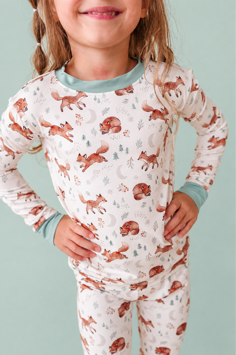 A young girl wearing a long-sleeve pajama set with an all-over woodland print of brown foxes, trees, and crescent moons on a white background, with light blue ribbed collar and cuffs. Bird and Bean Kids 